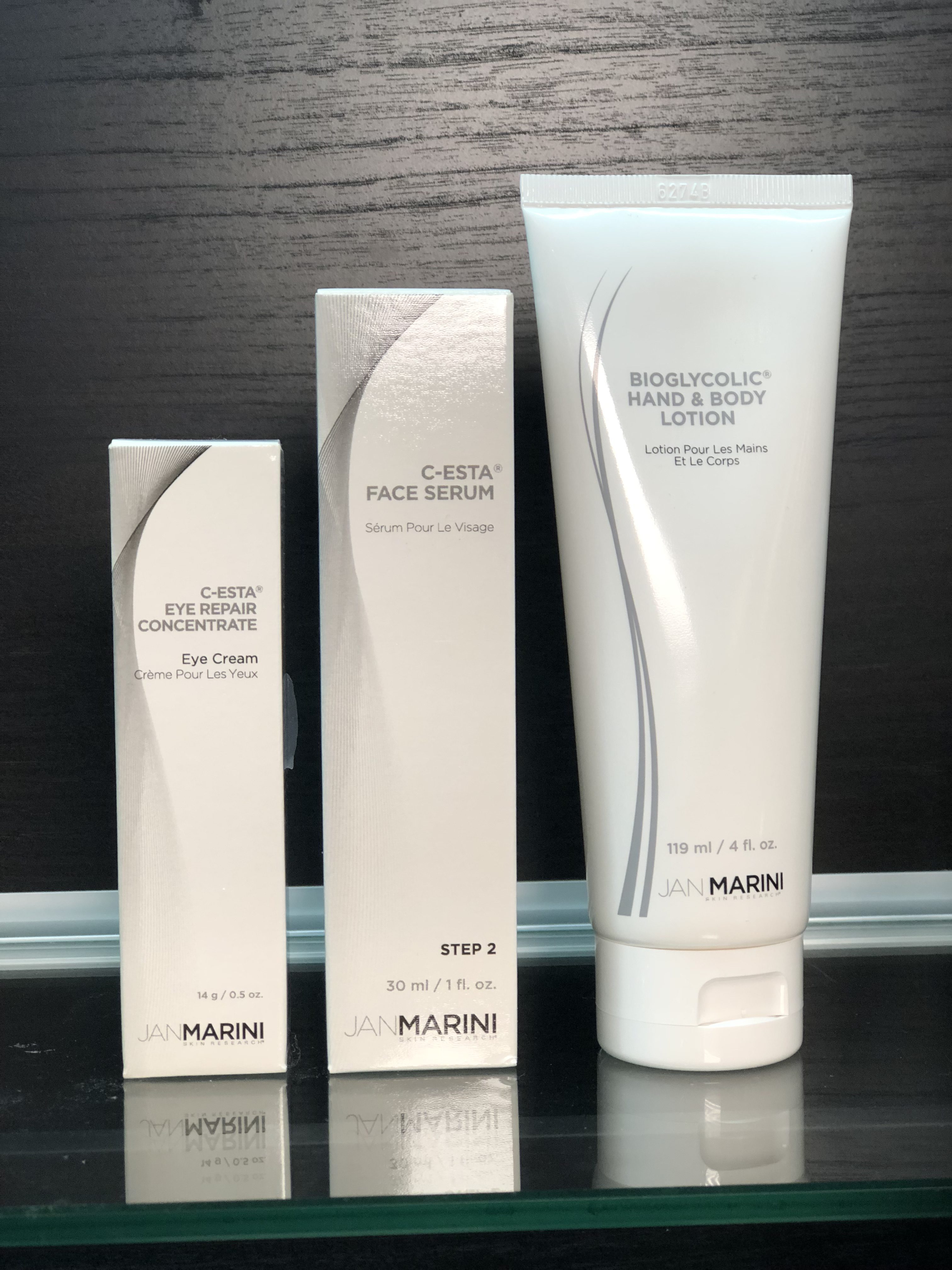 Image of 3 Jan Marini products side by side. From left to right: White box of C-Esta Eye Repair Concentrate, white box of C-Esta Face Serum, and white tube of Bioglycolic Hand & Body Lotion. All products are sitting on a glass shelf with a black wood textured background. 