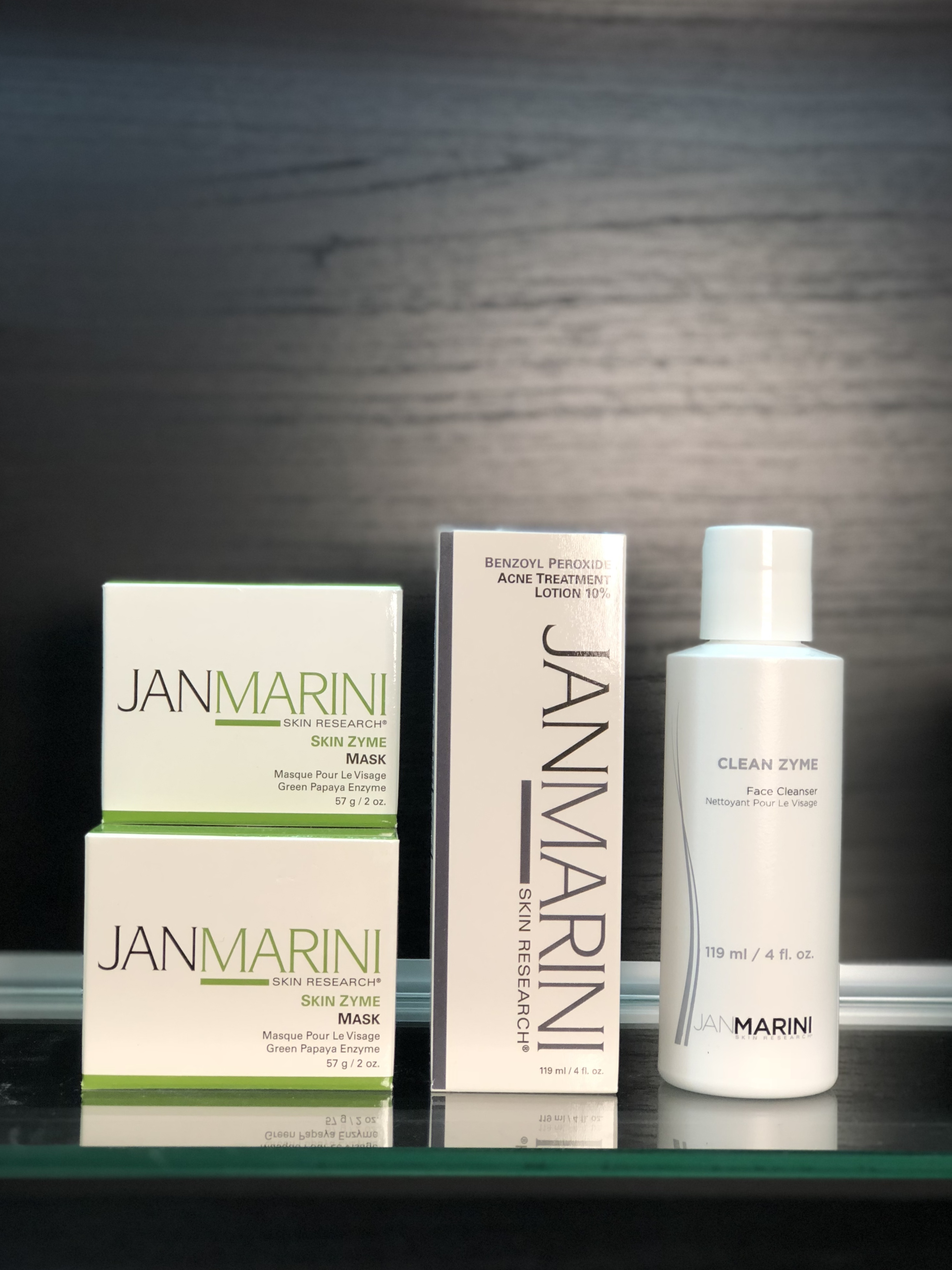 Image of 3 Jan Marini products. From left to right: two white boxes of Jan Marini Skin Zyme Mask stacked on top of each other, one white box of Benzoyl Peroxide Acne Treatment Lotion 10%, and one bottle of Clean Zyme Face Cleanser. All products are sitting on a glass shelf with a black wood textured background. 