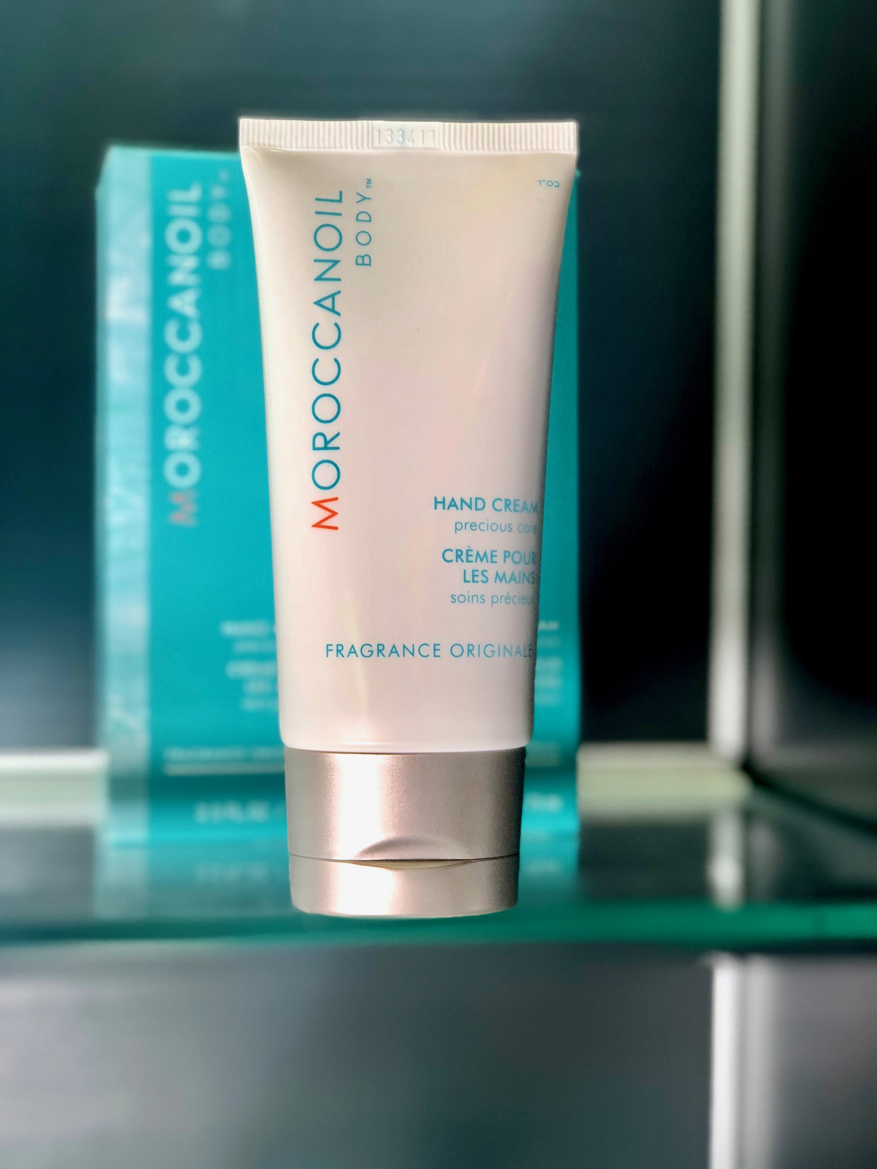 White tube with silver lid of Moroccanoil Hand Cream towards the front of a shelf and centered with two turquoise boxes of the same cream behind it slightly out of focus. 