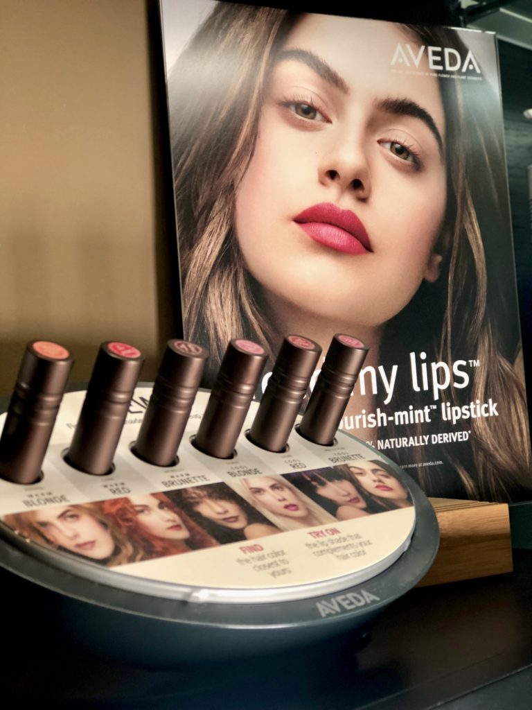 Image of display of 5 Aveda lipsticks aside promotional picture. From left to right: Lipstick display with five tubes in holders with photos of models underneath each one, and the Aveda logo at the bottom, photo of dark-haired model with pink lipstick, Aveda logo on the top righthand corner, and "Feed my lips" on bottom of picture. 